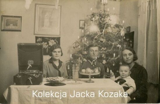 Na zdjęciu znajduje się żołnierz Korpusu Ochrony Pogranicza pozujący do zdjęcia z bliskimi. Siedzi pomiędzy dwiema kobietami, jedna z nich trzyma dziecko. W tle widoczna bożonarodzeniowa choinka.