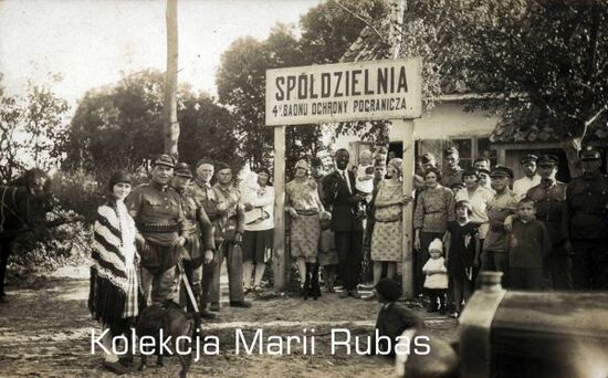 Zdjęcie pozowane grupy żołnierzy KOP wraz z rodzinami na tle Spółdzielni 4 Batalionu Ochrony Pogranicza.
