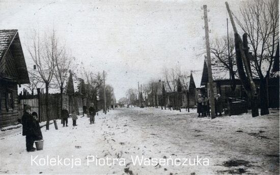 Zimowy widok ulicy w nieznanej miejscowości, szeregowa zabudowa po obu stronach drogi, ludzie stojący przed domami
