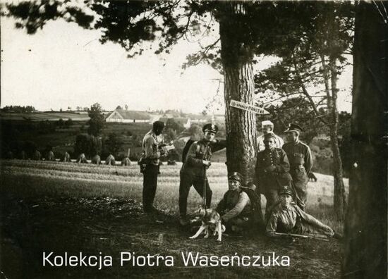 Grupa żołnierzy KOP pozująca przy drzewie z bronią i psem, na drzewie kierunkowskaz &quot;Do granicy państwa&quot;, w tle pole i zabudowania