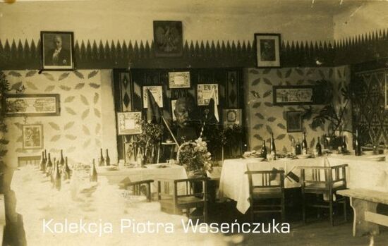 Przygotowanie do przyjęcia - zastawione stoły, na drugim planie popiersie Piłsudskiego z flagami i kwiatami, na ścianie godło oraz portrety prezydenta i marszałka